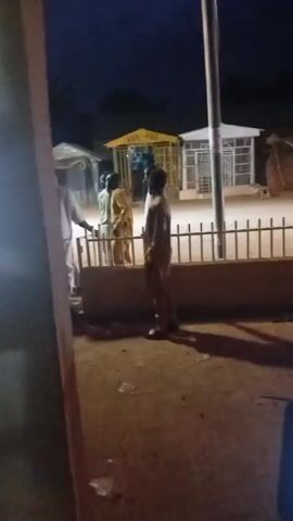 The video captures a nighttime scene outside with several people visible. In the foreground, a person holds a smartphone. Behind them, a fence separates the viewer from a street. Three individuals are standing near the fence, two of whom appear to be conversing. Beyond the fence, several small structures are visible, one with yellow trim and lettering that reads "PBS PBS". A vehicle with headlights on drives by, momentarily illuminating the area. Another vehicle appears to be parked or driving slowly behind the fence. The ground is dirt or pavement and debris is scattered. The lighting is dim, suggesting it is either dusk or night.
