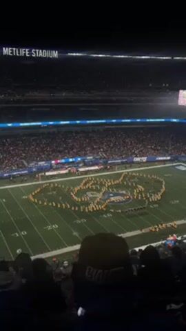 The video shows a marching band performing at MetLife Stadium. The stadium is packed with spectators. The marching band is on the football field and is arranged in a formation of the NY Giants logo. The band members are wearing yellow and black uniforms. The video is shot from a high angle, likely from the stands, and includes the back of the head of someone wearing a Giants hat.