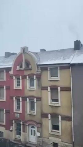 The video shows a view of several buildings during a light snowfall. The buildings are three stories tall and have different colors. The building on the left is painted in a pinkish red and has a yellow accent around the top window. The building in the middle is yellow with red accents and the building on the right is a neutral gray color. The roofs of the buildings are lightly covered in snow. Smoke rises from a chimney on the middle building. Snowflakes are visible falling in the foreground, indicating a wintry weather condition.