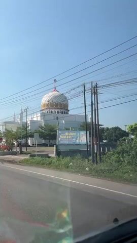 A white mosque with a distinctive gold and white dome, topped with a crescent moon symbol, is visible on the left side of the frame. The mosque is surrounded by trees and a low fence. A street runs in front of the mosque, with a car visible in the foreground, suggesting the footage is being filmed from inside a vehicle. There are power lines strung across the sky. A sign, partially obscured by trees, is visible on the right side of the road. The sky is clear and blue. A person on a motorcycle passes by on the street. The overall scene appears to be an urban or suburban environment.