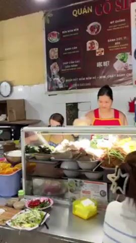 The video opens with a view inside a food stall, featuring a food display case with various ingredients. Two women stand behind the case preparing food. Customers are seated at tables, eating. The camera then pans to show a crowded indoor market. People are walking through the narrow aisles filled with stalls selling food and various goods. The market has a corrugated metal ceiling with fluorescent lighting.