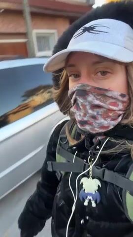 A masked person wearing a black jacket, white hat, and gloves with a skeleton print is seen outside. The person is standing on a colorful skateboard. The background features trees and a street with parked cars. The person raises their middle finger to the camera.