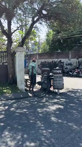 The video shows a person on a bicycle loaded with goods, standing on a paved street. The person, wearing a patterned shirt and dark pants, is facing away from the camera and appears to be stationary. The bicycle has two large baskets attached to the rear, filled with items. A large tree casts a shadow over the scene. In the background, a wall with graffiti is visible, as well as buildings and a utility pole. The overall environment appears to be an urban or semi-urban setting on a sunny day.