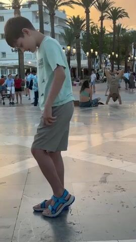 A boy in a light blue t-shirt and khaki shorts walks on a checkered tiled surface. He is wearing blue sandals. In the background, another person is doing a handstand. Palm trees and buildings are visible in the distance, along with other people walking around. The sky appears to be sunset colored.