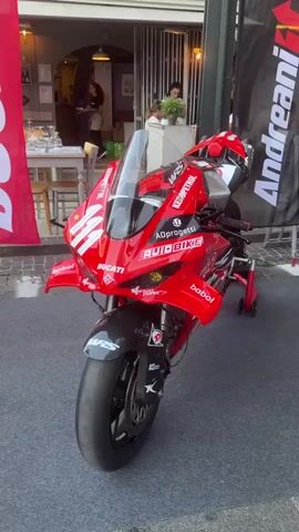 The video begins by showing the front and side of a red Ducati motorcycle with various sponsor decals. The motorcycle is displayed on a grey carpeted surface, likely for an exhibition or event. In the background, there are people seated at tables outside a building, suggesting a cafe or restaurant. A red banner with white lettering reading "DUCATI" is visible on the left side of the frame. As the video progresses, the camera pans to the right, showing the back of a person wearing a red shirt and blue denim shorts, standing next to another motorcycle. This second motorcycle is black with gold accents and is displayed on a table with the words "MOTO CORSE" visible. The environment appears to be an outdoor street setting, possibly a pedestrian area, with a building and more people in the background. The camera movement is fluid and pans from left to right, revealing different aspects of the display.
