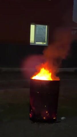 A video captures a fire burning inside a metal barrel at night. The barrel, dark and cylindrical, sits on a dirt or gravel surface, with small flames flickering from its base. The main blaze erupts from the top of the barrel, sending a thick plume of orange and yellow smoke into the dark sky. The fire casts a warm glow on the barrel's sides and illuminates the surrounding area with an eerie light.

In the background, a building with dark brickwork is visible. A single window on the building is illuminated from within, casting a pale yellow light onto the exterior. The setting appears to be outdoors, possibly a backyard or an open area near a residential building. The time of day is clearly night due to the darkness and the presence of artificial light from the window. The weather is not discernible from the video.

The activity is the burning of something within the barrel, likely for disposal or warmth. The scene is somewhat desolate, with the fire being the sole source of intense visual activity and warmth. There are no people visible in the video, and the focus is entirely on the burning barrel and its immediate surroundings. The overall mood is one of stark contrast between the intense, consuming fire and the quiet darkness of the night, creating a slightly dramatic and isolated atmosphere. No text is visible in the video. The location context provided is Şalqar, Kazakhstan.