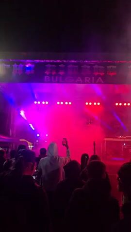 A crowd of people is silhouetted in front of a stage bathed in intense pink and purple lighting. A banner reading "HOODBOYZ BULGARIA" hangs above the stage. Several spotlights shine across the stage, obscured by fog or smoke. One person in the crowd is holding up a smartphone to record the scene. The stage is mostly empty save for a person in all black at the center behind a DJ setup.