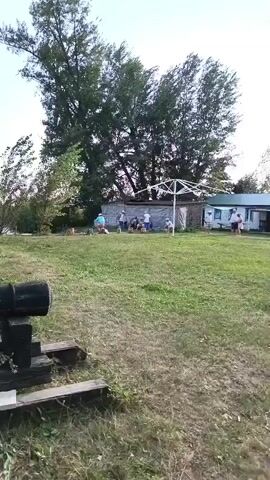 The video pans across a grassy outdoor area. In the foreground, a black cannon rests on a wooden carriage. Behind the cannon, several people are gathered around a white structure, possibly a tent or canopy. Further in the background, a tall wooden tower with a red flag atop it is visible. Trees and foliage surround the scene, and a body of water can be seen in the distance. The overall impression is that of a historical reenactment or a themed recreational area.