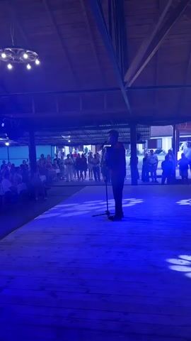 The media depicts a performance on an outdoor wooden stage illuminated by bright blue lights. A lone male figure, silhouetted against the lights, stands center stage holding a microphone and appears to be singing or speaking. He is dressed in dark clothing. The stage floor is also wooden and shows circular light projections.

In the background, an audience is seated at tables, observing the performance. The setting appears to be an open-air venue, possibly a pavilion or covered area, with a wooden roof structure visible overhead. Lights are suspended from the ceiling, including a decorative circular fixture. Beyond the audience, there are glimpses of buildings, suggesting an outdoor gathering or event.

The time of day is likely evening or night, given the artificial lighting and the overall ambiance. The event seems to be a concert, speech, or performance of some kind, with the audience actively engaged. The overall mood is festive and celebratory, indicated by the lighting and the presence of a gathered crowd. No visible text is present in the media.