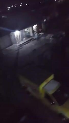 The video is shot at night, possibly from an elevated position. A yellow truck with a covered bed is visible. There is street lighting illuminating a courtyard area with some buildings around it. People are moving in the courtyard, although it's hard to discern details due to the darkness. The camera appears to be moving, creating a blurry effect as the viewer shifts perspective around the scene.