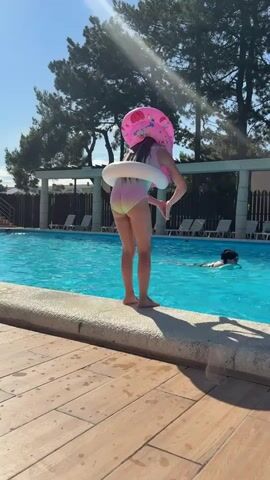 A young girl wearing a swimsuit, a pink inflatable ring, and an inflatable donut around her waist stands on the edge of a swimming pool, bending over. She is facing away from the camera, and her feet are positioned on the pool's edge. In the background, another person is swimming in the bright blue water. The sun is shining, casting a warm glow and creating visible light rays through the trees that line the perimeter of the pool area. The setting appears to be a resort or a public pool, with lounge chairs visible behind a fence. The overall mood seems to be one of fun and recreation on a sunny day. There is no visible text in the media file.