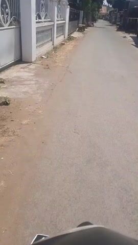The video shows a straight, paved road with houses and some trees lining both sides. The road appears to be in a residential area, likely in a warm climate due to the clear blue sky and bright sunlight. There are no people visible in the footage, but there are some objects that suggest human activity. On the left side of the road, a white fence with decorative metalwork encloses a property. A small Indonesian flag is visible on a flagpole in front of one of the houses. On the right side of the road, several small structures, possibly shops or storage units, are present, with clothes hanging out to dry in front of one. Wooden benches or tables are placed outside some of the buildings. The overall impression is of a quiet, ordinary street scene in a developing country.