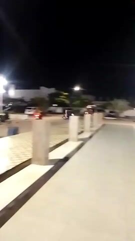 The video shows a blurred nighttime scene outdoors, possibly a parking lot or a public square. The camera is moving, creating a sense of motion. In the background, there are several vehicles, including cars and what appear to be motorcycles. Some lights are visible, illuminating the area. A few figures of people are also discernible, some sitting and others possibly standing or walking. The ground in the foreground has a patterned tile design. The overall atmosphere is indistinct due to the blur and low light conditions.