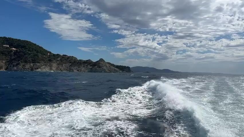 The video shows a view from a moving boat, looking towards a rugged, tree-covered island. The sea is a deep blue with white foam trailing behind the boat, indicating its speed. The sky is a mix of blue with scattered white clouds. The island has steep, rocky cliffs and appears to be sparsely populated, with a few buildings visible on a hilltop. The water is choppy, with waves breaking near the shore of the island. The horizon stretches out to the right, showing more islands in the distance. The overall impression is one of a scenic journey through a coastal landscape.