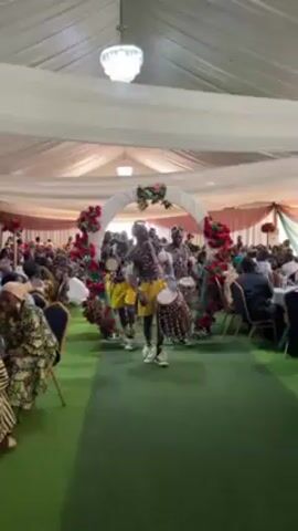 The video shows a group of African drummers performing at what appears to be a wedding reception. The reception is taking place in a large tented space with green carpet and white drapery overhead. Tables are set up for guests, who are seated and watching the performance. The drummers are wearing yellow shorts and traditional African attire including shell ornamentation and headbands. They are carrying and playing various sized drums, moving towards the camera. A decorated archway is visible behind them, adding to the festive atmosphere.