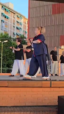 A group of people are performing what appears to be a martial arts demonstration on a raised stage. The person in the foreground, a woman with blonde hair pulled into a ponytail, is wearing a navy blue martial arts uniform with red trim and white sneakers. She is holding a long, thin stick, possibly a sword or staff, with both hands and appears to be in mid-movement. Behind her, several other individuals, dressed in black tops and white loose-fitting pants, are also holding similar sticks and moving in unison. The stage is made of wood and is supported by a brick structure. In the background, there is a large wooden wall with vertical red and natural wood colored slats, and beyond that, a building with multiple floors and balconies. The lighting suggests it is daytime, possibly late afternoon, given the shadows and the warm light. The overall atmosphere seems to be one of a public performance or demonstration.