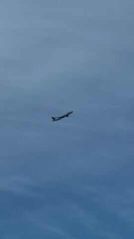 A plane is flying across the sky. It is a bright, clear day with a few wispy clouds. The plane is white with red accents on its tail. It is flying at a high altitude, and it appears to be a commercial airliner. The plane is moving from left to right across the frame, and it is ascending.