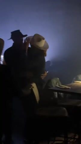 The video depicts a dimly lit scene with several individuals wearing cowboy hats, possibly at a bar or event. The subjects are mainly silhouetted against a blue background, which might be from stage lighting. A table and some bar stools are visible, suggesting a social gathering place.  The people are standing and seem to be interacting, with some movement suggesting dancing or celebration. The video is shot from a low angle, and the focus is mainly on the people in the hats.
