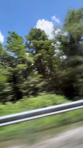 This is a first-person perspective video, likely filmed from a vehicle moving along a road. The scene shows lush green trees and foliage blurring past on the right side of the frame, indicating motion. A silver guardrail runs parallel to the road in the foreground, also exhibiting motion blur. Above the trees, a bright blue sky with scattered white clouds is visible. The overall impression is one of a scenic drive through a wooded area on a sunny day. There are no people or distinct objects clearly visible beyond the natural environment and the guardrail. The audio consists of a musical track.