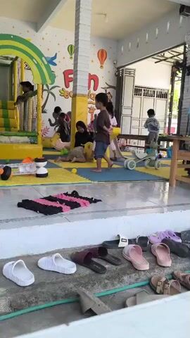 The video shows an indoor playground area for children. There are several children playing on various equipment. In the background, a large inflatable structure with a rainbow arch is visible. To the right of this, there are other play structures, including a yellow see-saw with two children on it, and a green balance bike. A woman is also present, interacting with some of the children. The floor is covered with colorful mats. In the foreground, a ledge is lined with a variety of children's and adult footwear, including several pairs of white slides, dark brown slides, pink slides, purple slides, and sandals. The overall environment appears to be a well-lit, child-friendly space.