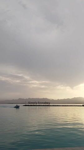 The scene is a daytime shot of the sea at "Simply Somabay", with mountains visible in the background. The sky is cloudy with a gradient of white and light gray. The water reflects the sky and has gentle ripples. A boat is moving from left to right across the water. There is a pier-like structure with signage that spells out "Simply Somabay". The light suggests either dawn or dusk.