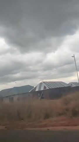 The video shows a view from a moving vehicle, likely a car, traveling along a road. The sky is overcast with heavy, dark clouds, suggesting impending rain or a storm. The landscape consists of dry, dusty ground in the foreground, with patches of vegetation and some buildings in the background. There are several structures, some appearing to be houses with corrugated metal roofs. Trees and tall grass are scattered throughout the scene. Power lines and utility poles are visible overhead, indicating the presence of infrastructure. The overall impression is of a rural or semi-rural environment experiencing inclement weather. The video captures a sense of movement and a slightly blurred view of the surroundings due to the motion of the vehicle and possibly wind.