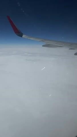 The video shows an aerial view from an airplane window. An airplane wing, with a red tip, extends into the frame from the top left corner, angled downwards and to the right. Below the wing, a vast expanse of white clouds stretches across the frame. The sky above the clouds is a deep blue. There are faint, bright white streaks visible within the clouds, possibly reflections or sunlight. The video appears to be a short, static shot or has very minimal movement.