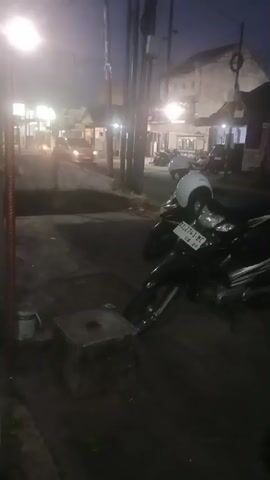 This is a vertical, low-light, handheld video showing a street scene at dusk. In the foreground, a concrete block sits on a sidewalk. To the right, two motorcycles are parked, one with a white helmet on its seat. The street beyond is dimly lit by streetlights and the headlights of a few passing cars. Buildings line the street. The overall impression is that of a quiet, everyday street scene in the early evening.