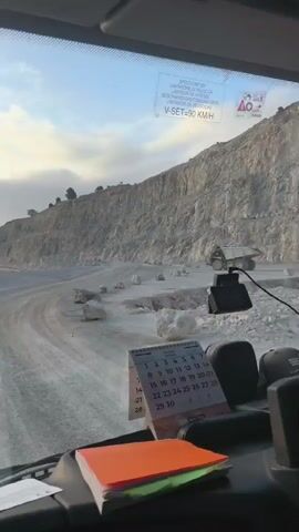 The video is taken from inside a vehicle, looking out through the windshield. In the foreground, on the dashboard, is a calendar for the month of October. To the right of the calendar, an orange notebook and a green notebook are stacked. The vehicle appears to be moving along a dirt road in a quarry. A large, steep, grey rock face dominates the landscape to the right, with a road curving around its base. To the left, the terrain slopes downwards, also covered in dust and small rocks. In the distance, a large dump truck with its bed raised is visible on the road. The sky is partly cloudy with a pale blue hue. The overall impression is a working quarry environment under a clear sky.
