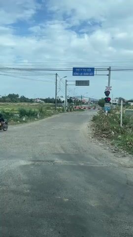 The video shows a rural road leading to a railroad crossing. The sky is cloudy with patches of blue. There are power lines overhead. A railroad crossing gate is down, indicating an approaching train. Red lights are flashing on the traffic signal, and a sign reads "CHÚ Ý TÀU HỎA DỪNG LẠI QUAN SÁT," which translates to "Attention Train Stop and Observe." There are also white stripes painted on the road, indicating a pedestrian crossing. The surrounding area is characterized by fields, some vegetation, and a few buildings in the distance. The road surface is asphalt and appears to be in fair condition with some cracks and potholes. The scene is static, and no people or vehicles are actively engaged in movement at the crossing itself.