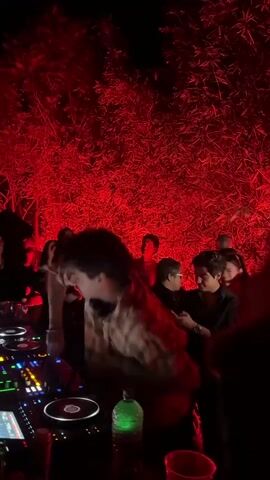 A DJ, wearing a plaid shirt and headphones around his neck, stands in front of a set of DJ equipment in a dimly lit outdoor space. Several people are gathered around him, some with their arms raised in the air, moving to the music. The background is illuminated with red lighting, creating a saturated, party atmosphere. A green bottle is visible next to the DJ equipment.