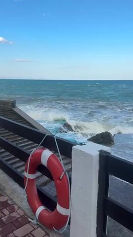 A life preserver hangs on a railing overlooking a staircase leading down to the sea. Waves crash against the shore and the stairs. The sky is blue with a few white clouds.