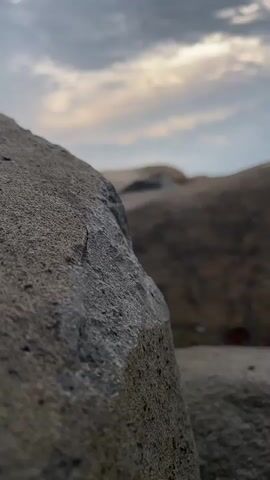 The video shows a close-up, low-angle view of rocks in the foreground, with a blurred background of more rocks and the sky. The sky is cloudy with patches of light, suggesting either dawn or dusk. The rocks are varied in size and texture, with the closest rock being large and rough. There are no people or animals visible in the video, and no actions or interactions are depicted. The environment appears to be a rocky outdoor area, possibly a beach or a mountainside.