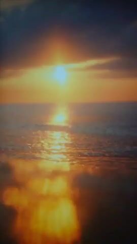 This is a video of a sunset over a body of water. The sun is low on the horizon, casting a bright orange and yellow light across the sky and reflecting on the water. The water is choppy, with small waves breaking on the surface. Dark clouds are visible in the upper part of the frame, contrasting with the bright colors of the sunset. The overall impression is one of a serene and beautiful natural scene.