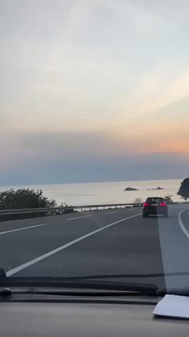 The video shows a car driving along a coastal road during a sunset. The sky is a mix of light blue, pink, and orange hues. The sea is calm, with a few small islands visible in the distance. Another car is driving ahead in the same lane. The road is asphalt with white lines marking the lanes. There is a metal guardrail on the left side of the road, separating it from the sea view. The view is from inside the car, looking out through the windshield.