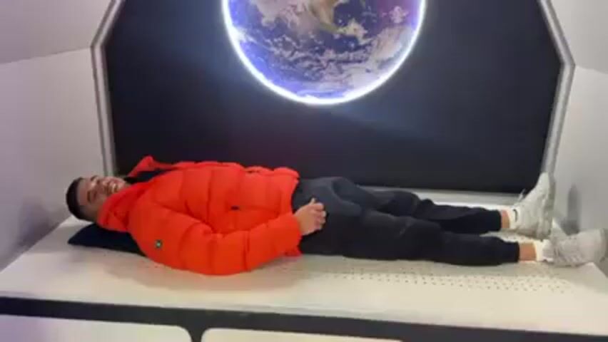 A young man, wearing a red jacket, black pants, and white sneakers, lies on a bed of nails. He is smiling and gives a thumbs-up. Behind him is a screen showing an image of Earth. The room appears to be a space-themed exhibit or display.