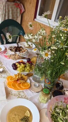 The video shows a table laden with food and flowers, likely set for a meal outdoors. In the foreground, a white plate holds a portion of what appears to be lentil soup with a slice of lemon and some herbs. To the right, a pink bowl contains a salad of shredded cabbage, cucumbers, and herbs. Beyond this, a plate holds a portion of grain with chopped tomatoes. Several other plates are visible, some with similar portions of grain and tomatoes. A bowl of fruit includes grapes, plums, and apricots. A basket of bread sits in the background. A large bouquet of daisies and other small white flowers is prominently displayed in a glass vase. A small jar of honey with a green label is also on the table, as are a few small candles. A person's arm, adorned with bracelets, is visible reaching towards the table to adjust a plate. The setting appears to be a porch or patio, with wooden walls and a window visible in the background. A green plastic chair is also present.
