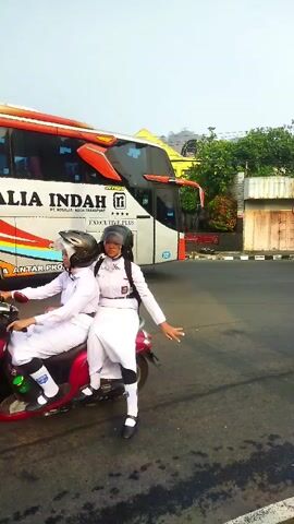 The video shows two young women in school uniforms riding a scooter on a paved road. They are both wearing helmets and white uniforms with red accents. The woman in the front is driving the scooter, and the woman in the back is looking at the camera and smiling. Behind them, a large bus with the text "TRANS INDAH" is visible. The environment appears to be a street with buildings and trees in the background. The lighting suggests it is daytime, likely morning or afternoon.