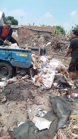 Two men are working at a garbage dump. One man, wearing a red vest and a green hat, is on the back of a blue truck, shoveling trash into the truck bed. The other man, wearing a black shirt and dark shorts, is standing in the trash pile next to the truck, using a long stick to push garbage towards the truck. The truck is filled with a variety of trash, including plastic bags, wrappers, and what appears to be organic waste. The ground around the truck is covered in mud and debris. In the background, there are some houses and a large pile of trash. The sky is blue with some clouds.
