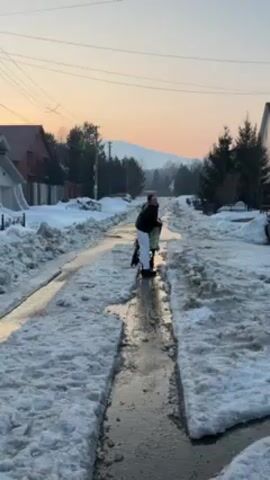 A person wearing a black jacket and white pants walks away from the camera, pushing a stroller down a narrow, icy road. The road is flanked by snowdrifts, and the setting sun casts a warm glow on the horizon, illuminating distant mountains. Houses and trees line the sides of the road, adding to the wintery landscape.