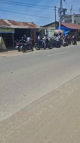 The video shows a roadside scene on a sunny day. A group of people on motorbikes are gathered on the side of the road, near a small shop with a rusty metal roof. Several individuals on motorbikes are driving on the road. The road is paved and there are utility poles visible in the background.  A building with a gray roof is also visible in the background.