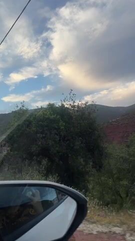 The view from a car traveling on a road shows a landscape of rolling hills and trees under a sky with scattered clouds. The hills in the distance have a reddish hue, contrasting with the green vegetation closer to the road. Some houses are visible among the trees. The car's side mirror is visible in the foreground, reflecting the interior of the car. Overhead electrical lines run parallel to the road.