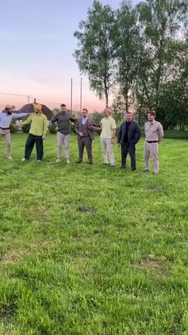The video shows seven men in a grassy yard. They are all dressed in different outfits. One man stands out in front and does a running split jump. The rest of the group react with laughter. One man on the right appears to film the stunt. Tall trees are in the background.