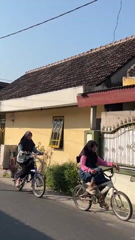 Two young women wearing hijabs ride bicycles down a paved street. The woman on the left is on a blue bicycle with a basket on the front. The woman on the right is on a silver bicycle with a pink skirt. They are riding past a yellow house with a tiled roof. The sky is clear and blue.