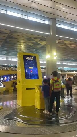 The video shows an indoor scene with a bright yellow arcade game. Two boys are playing the game, which appears to be called "Butter Stack." The game has a screen displaying colorful graphics and a red ball. In the background, other people are visible, and the environment appears to be a public space, possibly an airport or a mall, with a modern ceiling design.