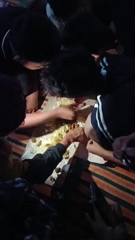 The video shows a group of young men eating together from a large spread of food laid out on paper on the ground. They are all leaning in towards the food, eating with their hands. The lighting is dim, suggesting it is nighttime or indoors with low light. The men are wearing casual clothing. The food appears to be a mix of rice and some sort of savory dish with vegetables. The atmosphere seems informal and communal.
