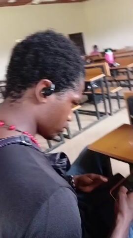 In a classroom setting in Gwafan, Nigeria, a young Black person is seated at a desk, engrossed in their mobile devices. The individual has short, curly black hair and is wearing a black t-shirt and blue jeans. They have a single black earbud in their left ear and a beaded necklace around their neck. Their attention is split between two smartphones; one is held in their right hand, displaying a grid of app icons, and the other rests on their lap, being manipulated with their left hand. The phone in their right hand appears to be an orange-cased smartphone.

The classroom is filled with rows of wooden desks with attached seating, typical of a school environment. The desks are simple and functional, with worn wooden surfaces. Sunlight filters in, suggesting it is daytime, though no specific time can be determined. The overall atmosphere appears to be one of quiet concentration or perhaps disengagement from a lesson, as the individual focuses on their phones. The presence of multiple desks in the background indicates that this is a shared learning space, but the focus is entirely on the individual and their technological interaction. There is no visible text on the objects or in the background that can be discerned. The scene is devoid of any overt emotional expression, portraying a moment of personal activity within a communal setting.