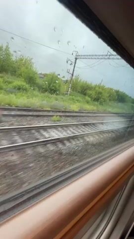 This video shows a view from a train window as it travels along railroad tracks. The window has raindrops on it, obscuring some of the view. Outside the window, there are multiple sets of railroad tracks, a grassy hillside with trees and bushes, and overhead power lines with support poles. The train appears to be moving at a moderate speed, indicated by the blurred motion of the tracks and landscape. The sky is overcast and appears to be a cloudy or rainy day.