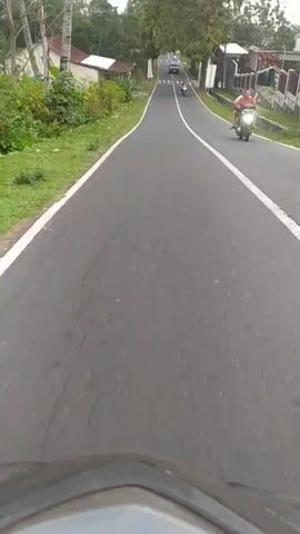 The video is a first-person perspective recording of riding on a road. The road is paved with asphalt and has a white line marking the right edge. There are trees and vegetation along the side of the road, as well as houses. Several vehicles are visible on the road, including motorcycles, a car, and a sidecar motorcycle. The rider appears to be moving at a moderate speed. The scene is rural or suburban. The recording is continuous and appears to be from a handheld device or a camera mounted on the vehicle.