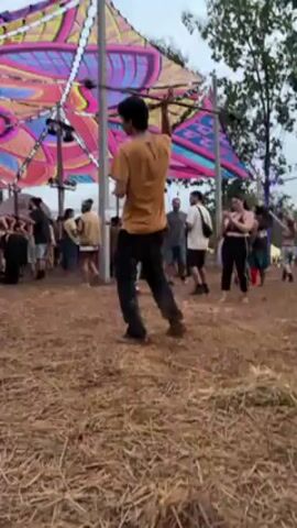 The video shows a crowd of people dancing under a large, colorful canopy supported by metal posts. The canopy has an intricate geometric design featuring vibrant shades of pink, blue, and yellow. A man in a yellow shirt and dark pants is energetically dancing in the foreground, raising his arms and moving rhythmically. Other people are seen scattered around, some dancing, some walking, and some standing still. A large black speaker sits near the edge of the canopy. The ground is covered in straw or wood chips, and trees are visible in the background, indicating an outdoor setting. The sky appears overcast.